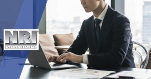 A male of unknown ethnic background is wearing a black suit blazer with dark blue tie and a white button down shirt. He is sitting at a desk and typing on a laptop, the image is zoomed in and his full face is not shown.