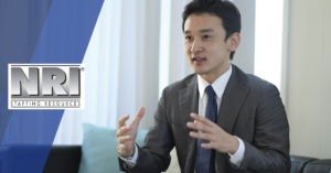 A male of Asian decent is sitting down in conversation, he is wearing a grey blazer with a white button down shirt and a blue tie