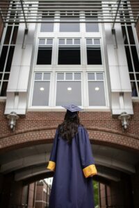 Recent college graduate in cap and gown facing campus building, considering first job out of college staffing agency options.